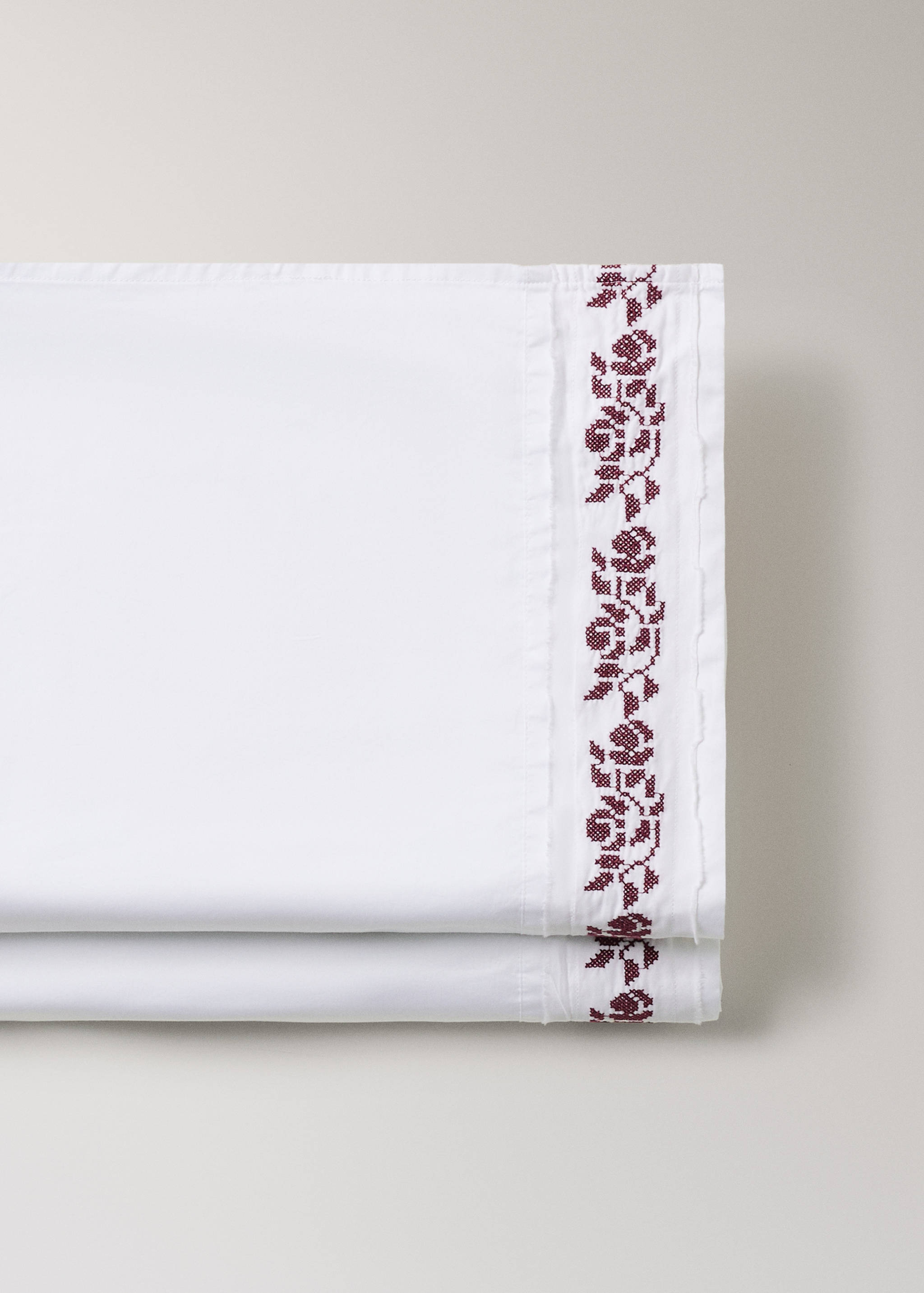 White flat sheet detail with burgundy cross-stitch floral embroidery.