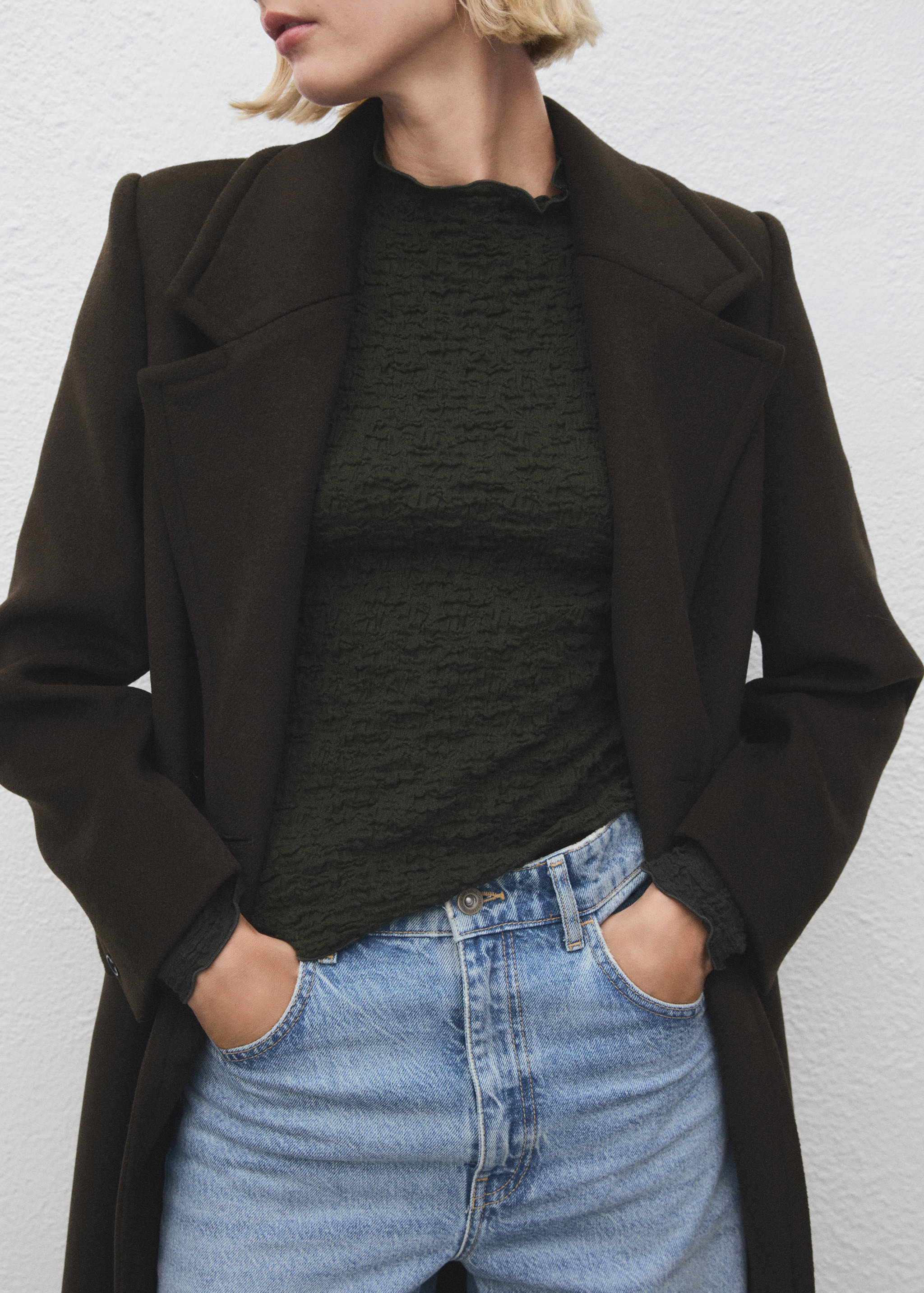 Dark green textured top with frilled mock neck, layered with a dark brown coat and blue jeans.