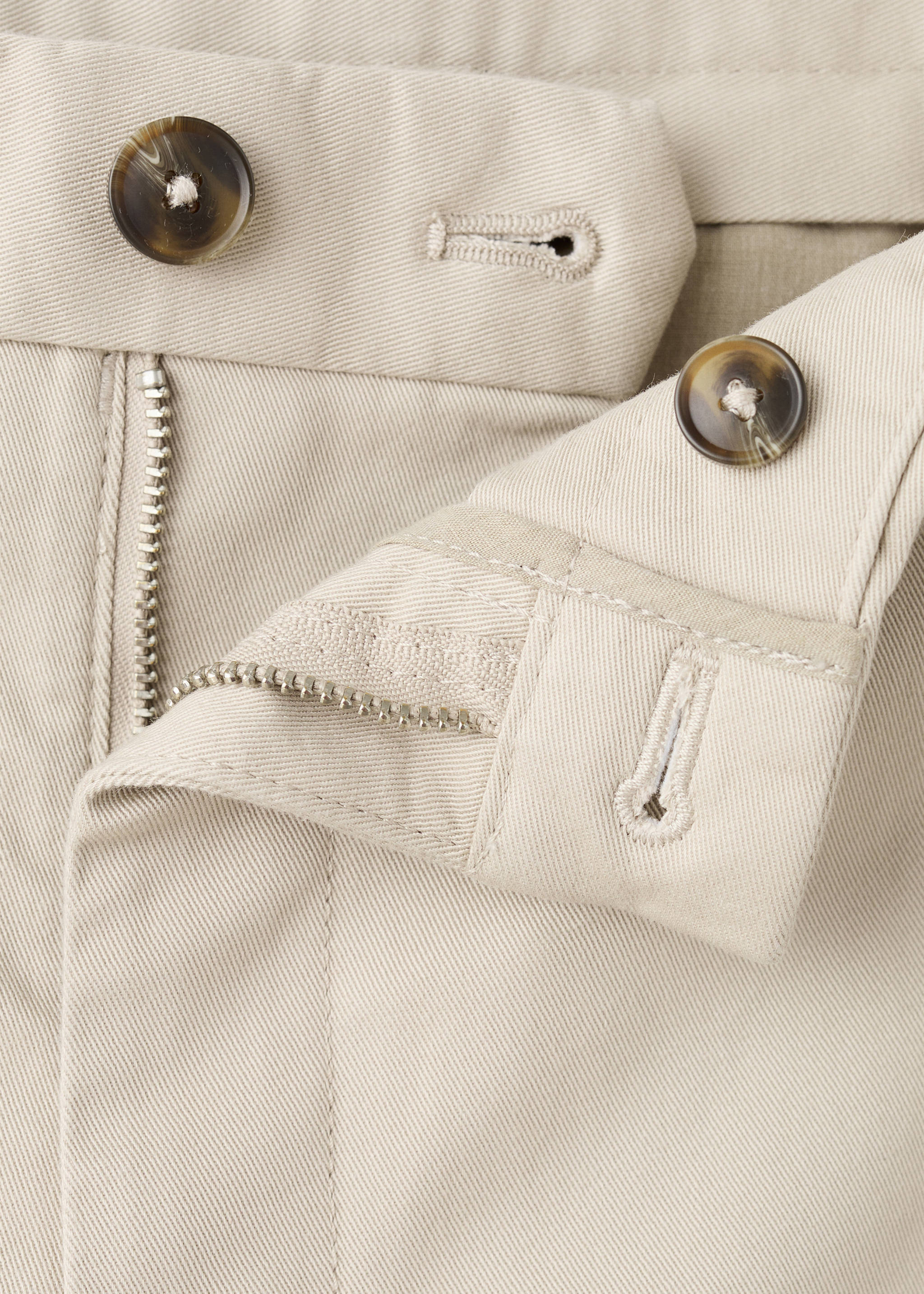 Beige trousers detail showing the waistband, tortoiseshell buttons, and zipper fly.