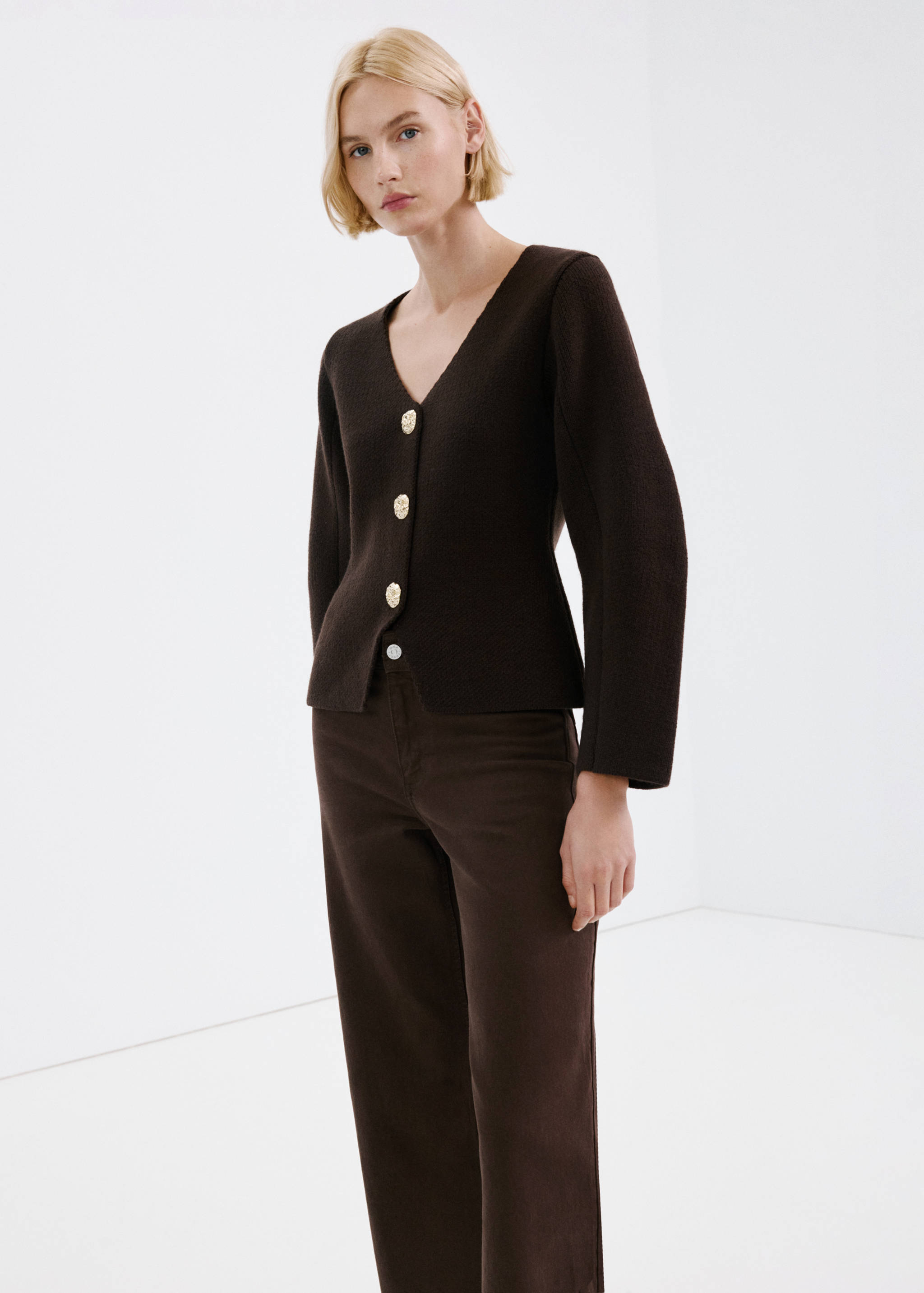 Dark brown knitted cardigan with V-neck and ornate gold buttons, worn with brown trousers.