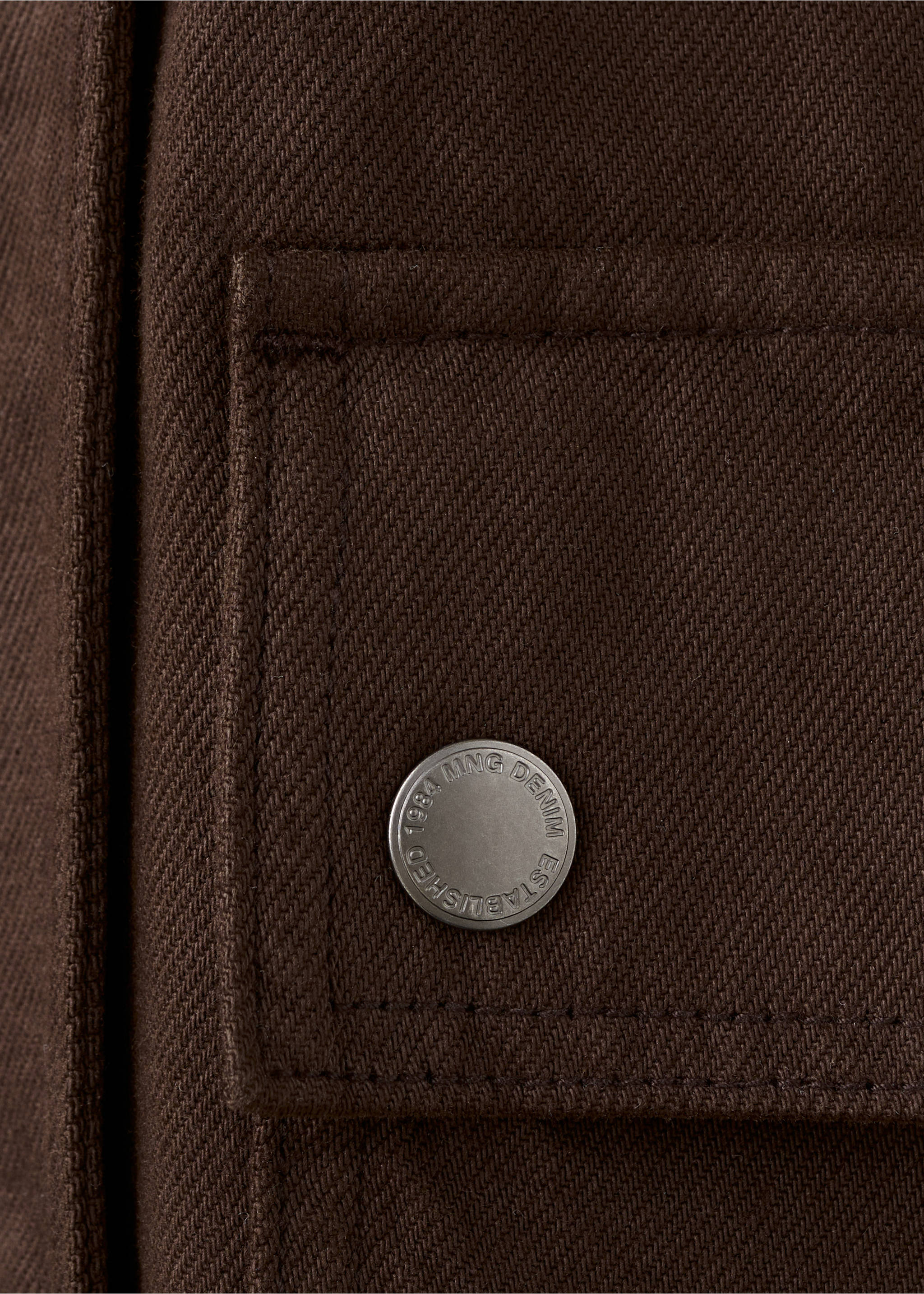 Close-up of a brown jacket pocket detail with an embossed metallic button., Chocolate. Ref: 27045819-00.