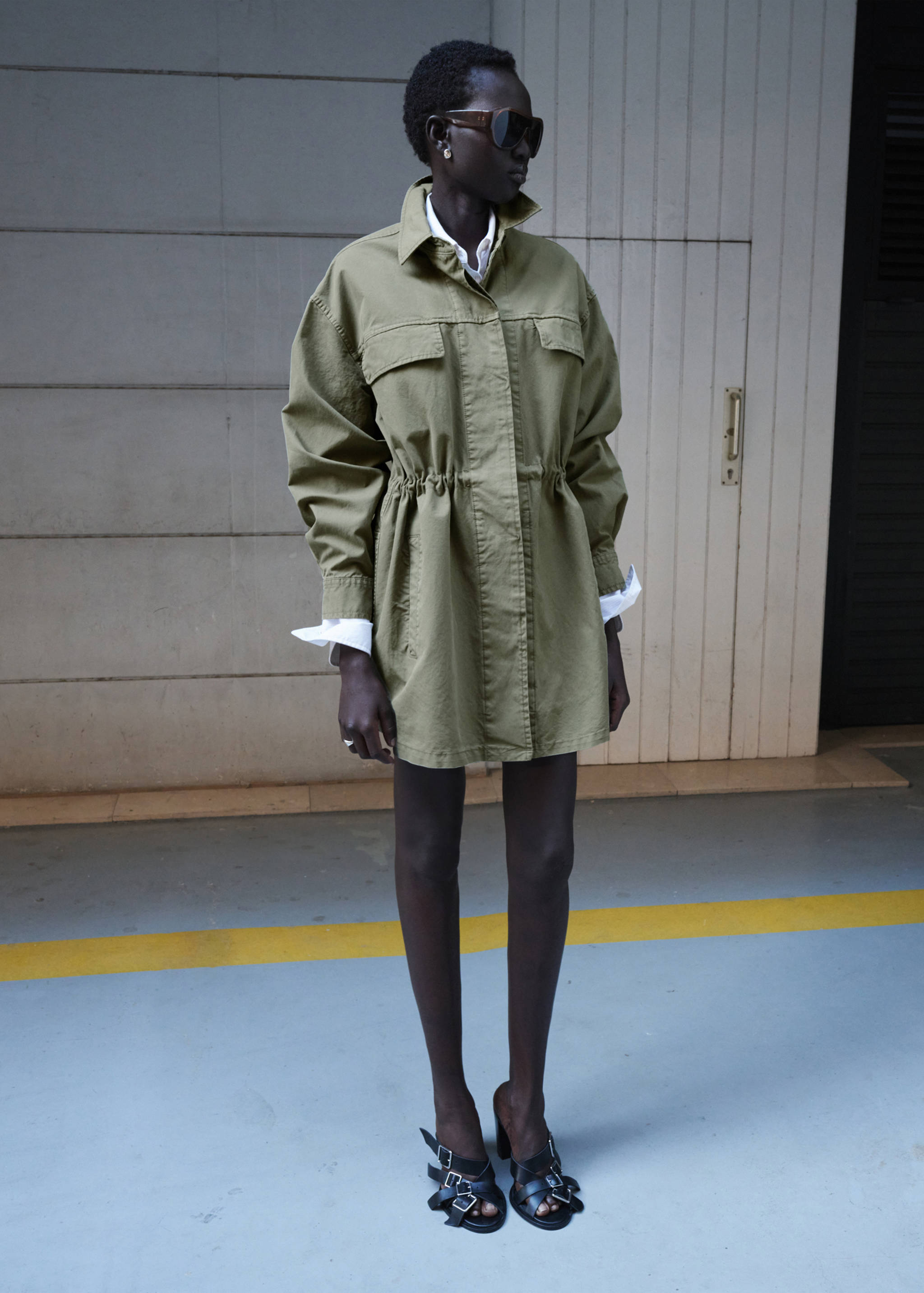Parka verde caqui con cintura ajustada y bolsillos de solapa en el pecho, combinada con una camisa blanca.