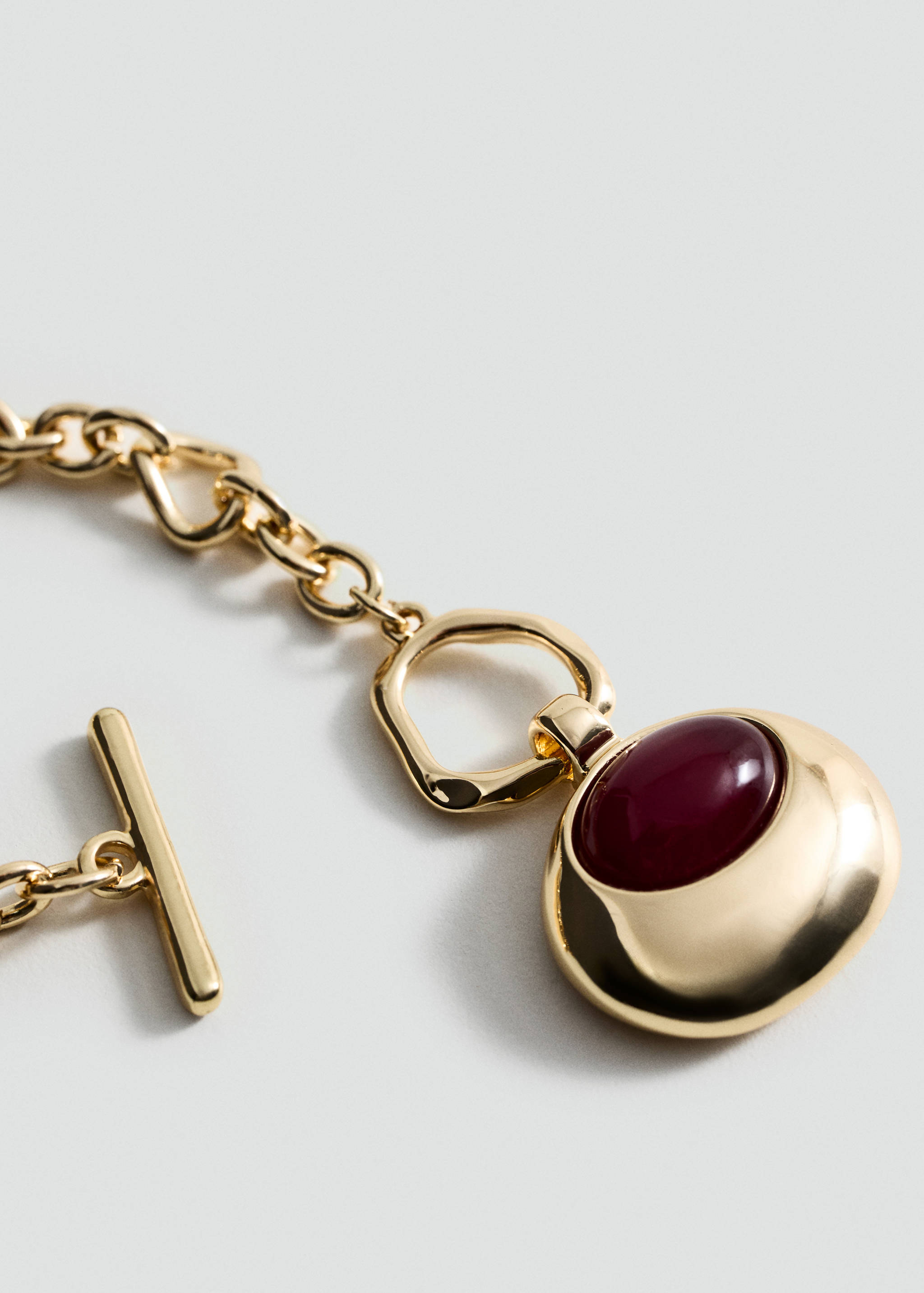 Gold-toned metal collar necklace with a chain link, T-bar clasp, and a pendant with a red cabochon stone.