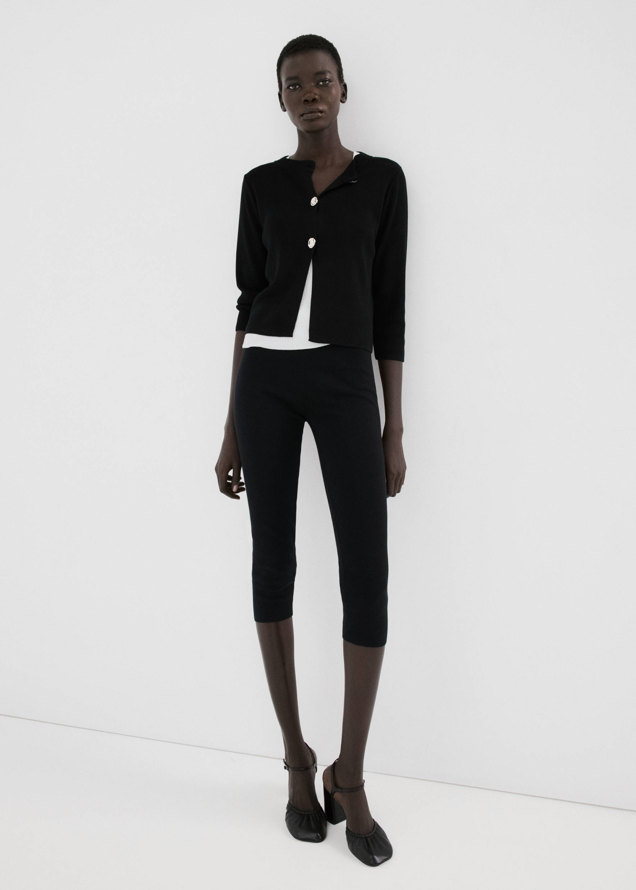 Black cardigan with jewelled buttons worn over a white top with black trousers.