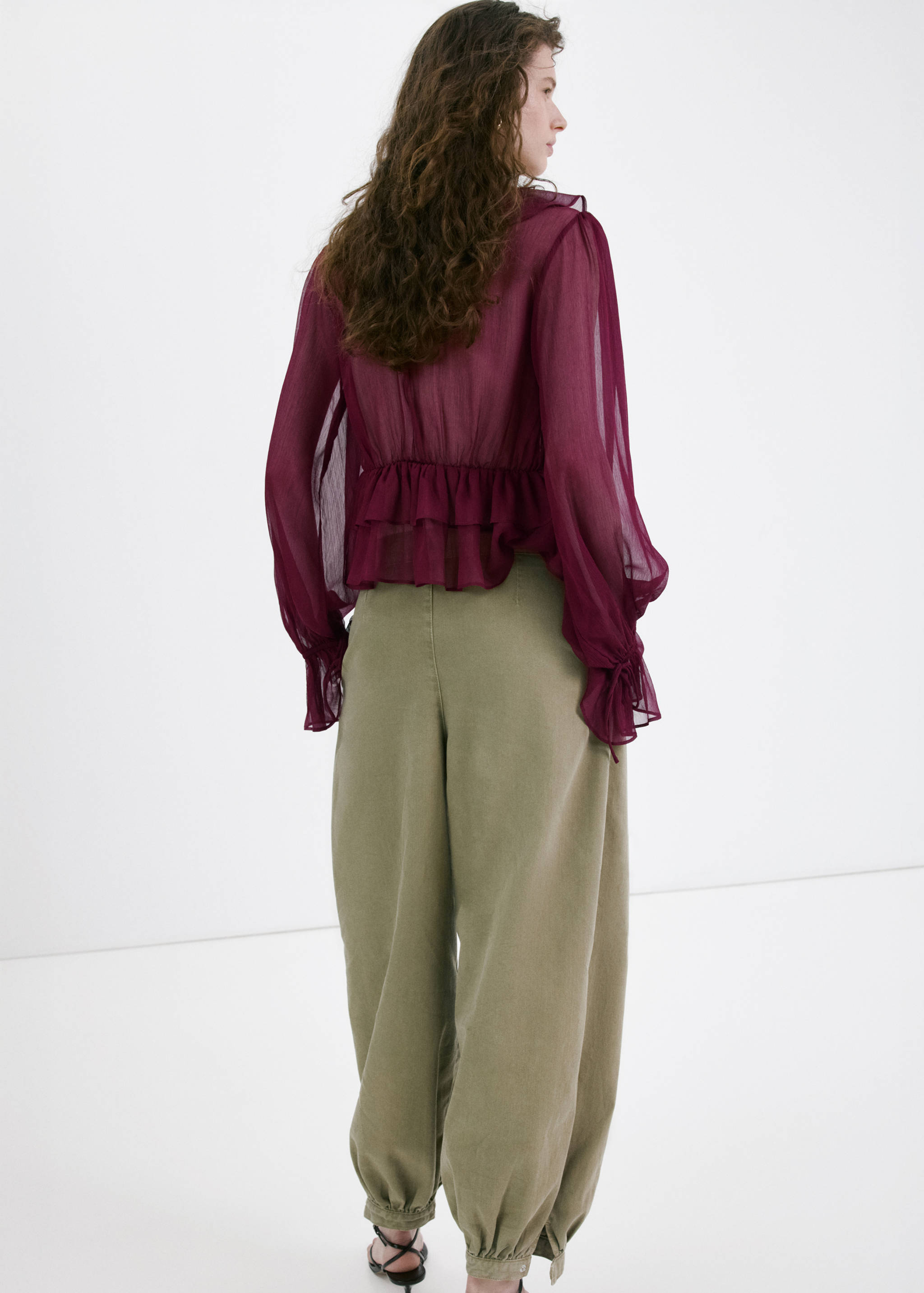 Back view of a plum sheer blouse with ruffled hem, long sleeves, and olive green trousers.