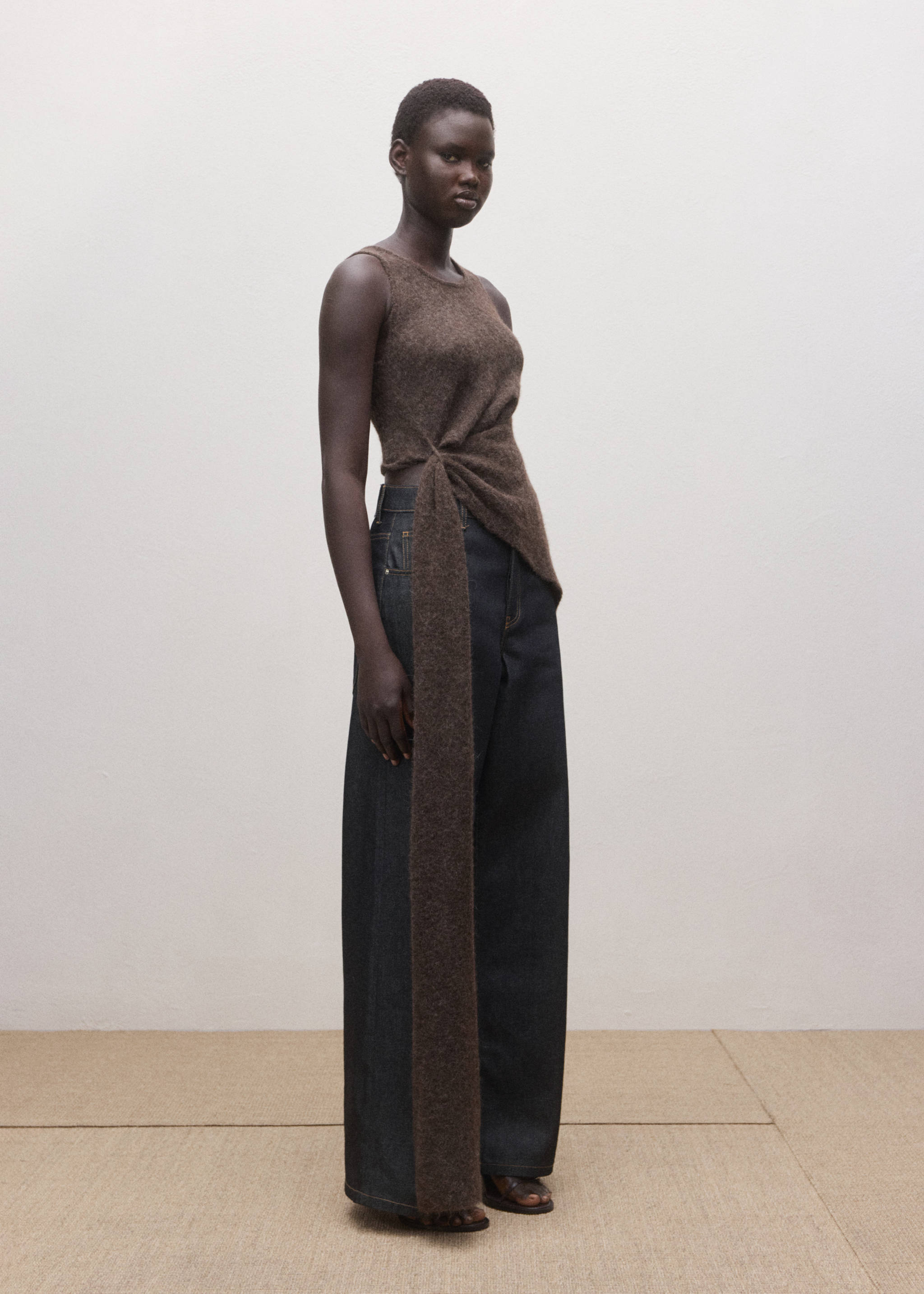 Dark brown sleeveless knitted top with a side knot and long sash, paired with wide-leg trousers.