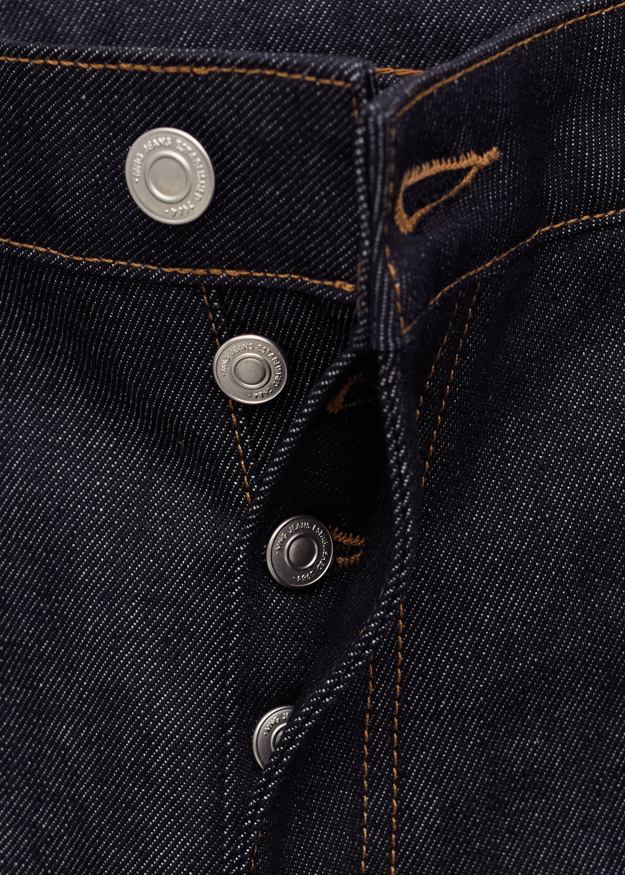 Close-up of dark blue denim trousers showing the metallic button fly and contrast topstitching.