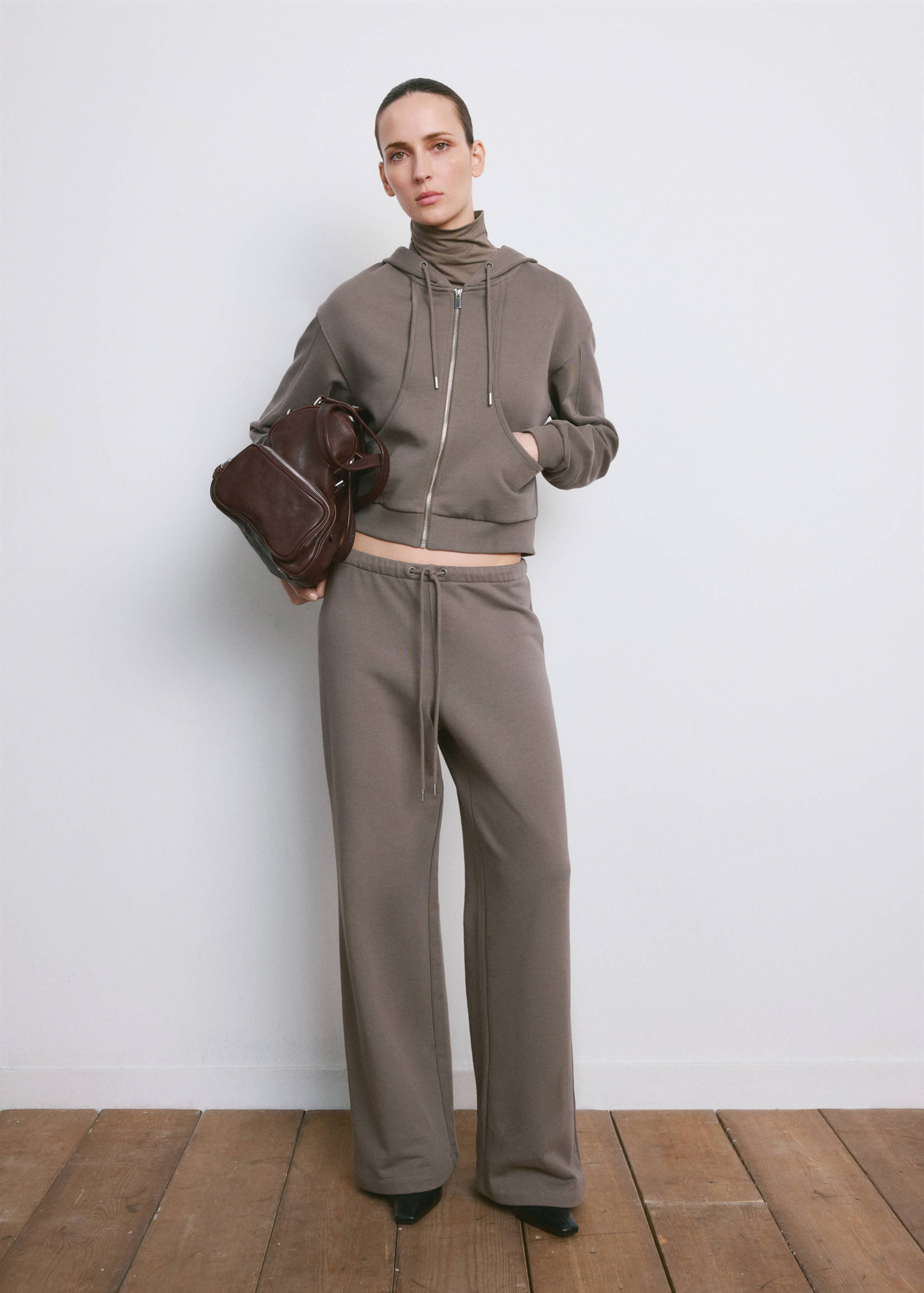 Grey-brown cropped zip hoodie with drawstring, worn with matching trousers and a dark brown bag.