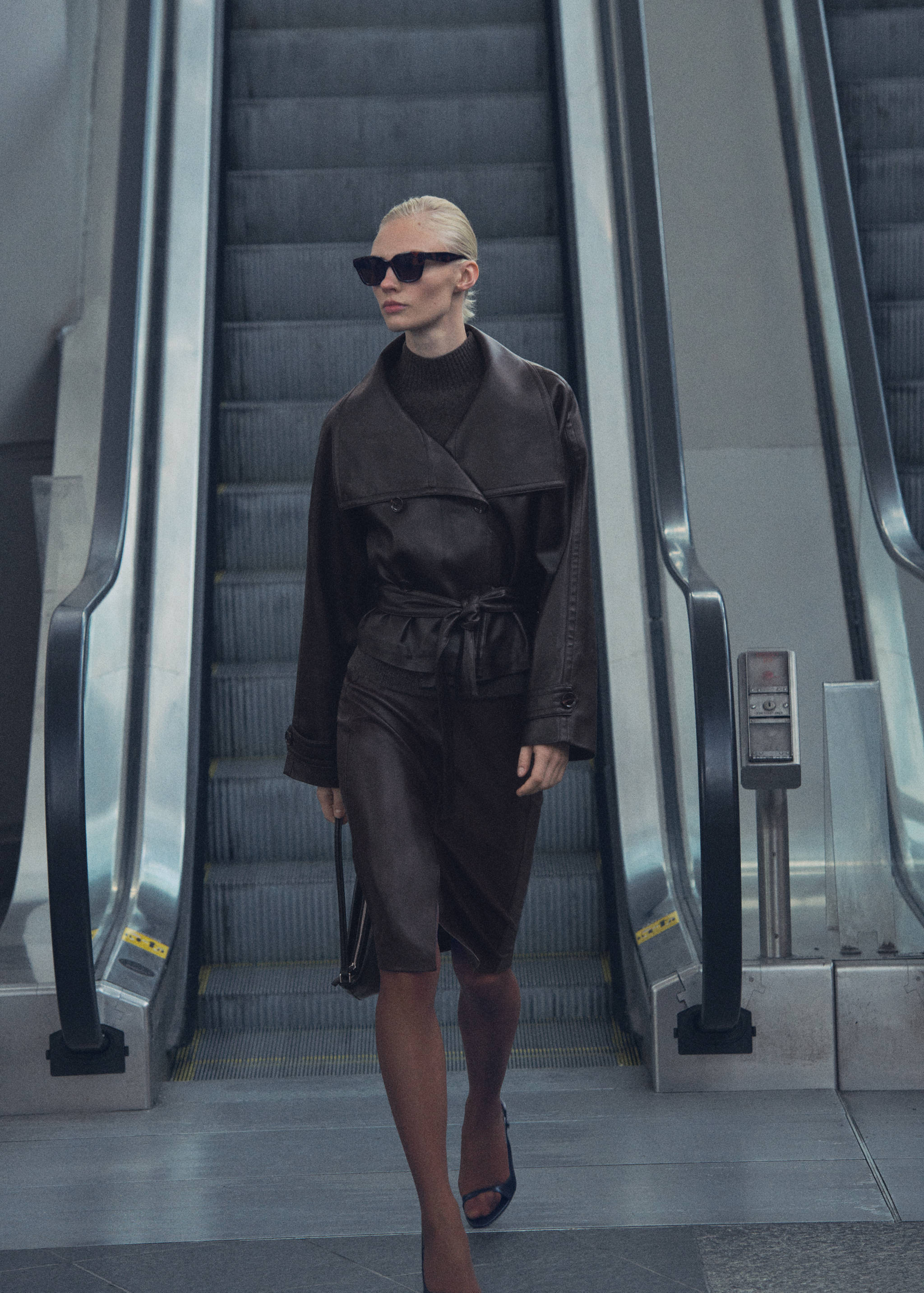 Dark brown leather jacket with an oversized collar and tie belt, worn descending an escalator.